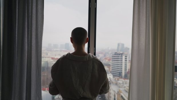 young-unhappy-woman-with-cancer-standing-near-a-window-and-looking-at-view-SBI-351324725